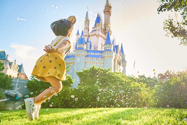 A girl with gold Mickey ears jumping through bubbles with Cinderella Castle in the background.